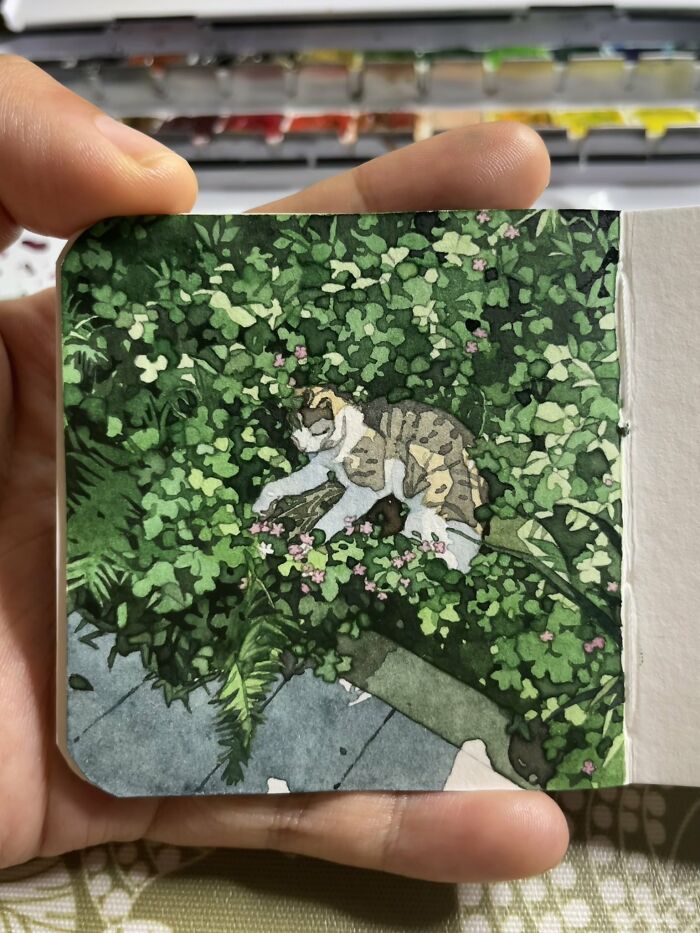 Hand holding a small watercolor painting of a cat resting among green foliage and pink flowers in a dreamy style.