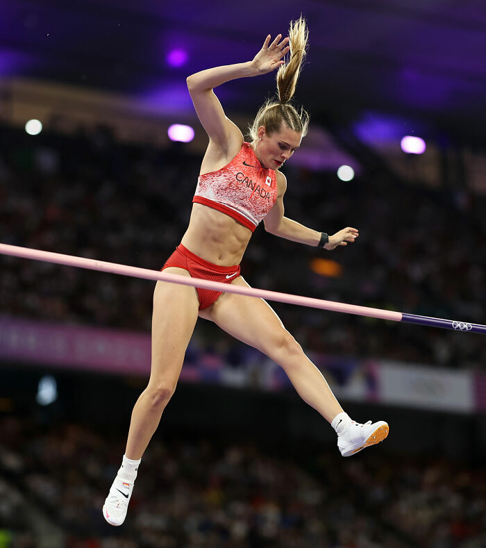 Olympic pole vaulter Alysha Newman competing mid-air wearing red Canada uniform during a pole vault event at a major athletics competition.