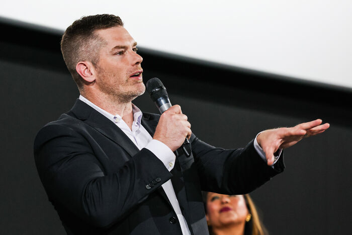 Man in a black suit speaking into a microphone, discussing The Chosen season 6 release date and season 7 update.