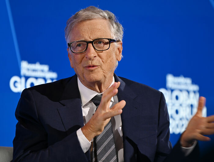 Bill Gates speaking at a conference, wearing glasses and a suit, addressing Epstein files and related accusations.