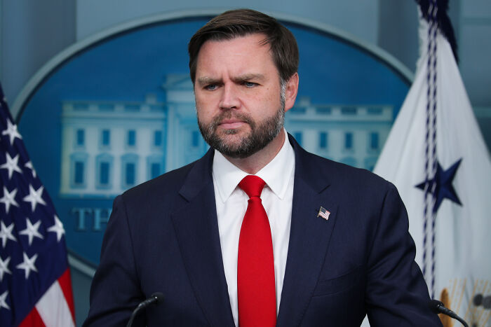 JD Vance speaking at a podium with American flags in the background, addressing comments on Eileen Gu’s representation.