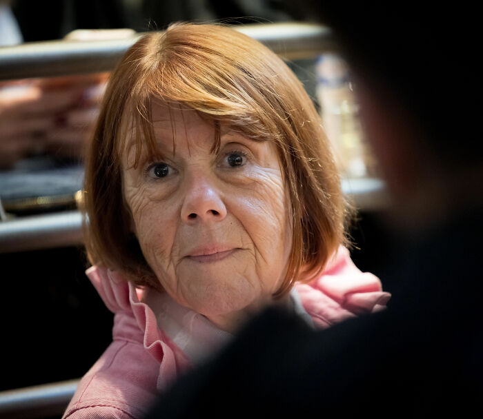 Gisèle Pelicot with reddish hair, wearing a pink shirt, showing a serious expression during a candid indoor moment.