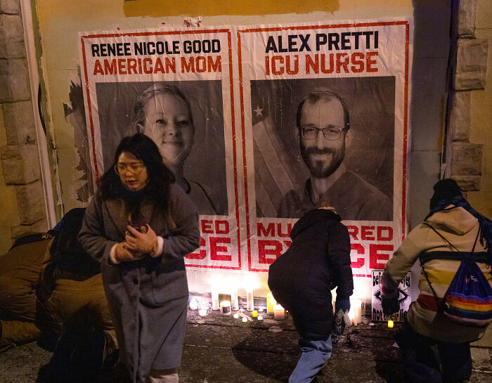 People lighting candles at a nighttime vigil in front of posters of Alex Pretti, ICU nurse, and Renee Nicole Good, American mom.