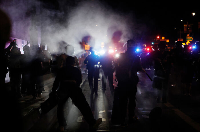 Police officers stand amid smoke and flashing lights during a tense night scene related to Alex Pretti's case update.