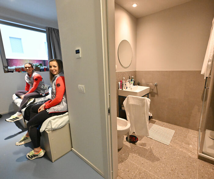 Two athletes sitting in a Winter Olympics Milan room, showing the compact space and simple bathroom setup.