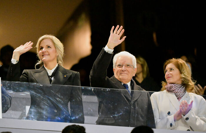 Winter Olympics host waving and smiling with two people in formal attire during a public event.