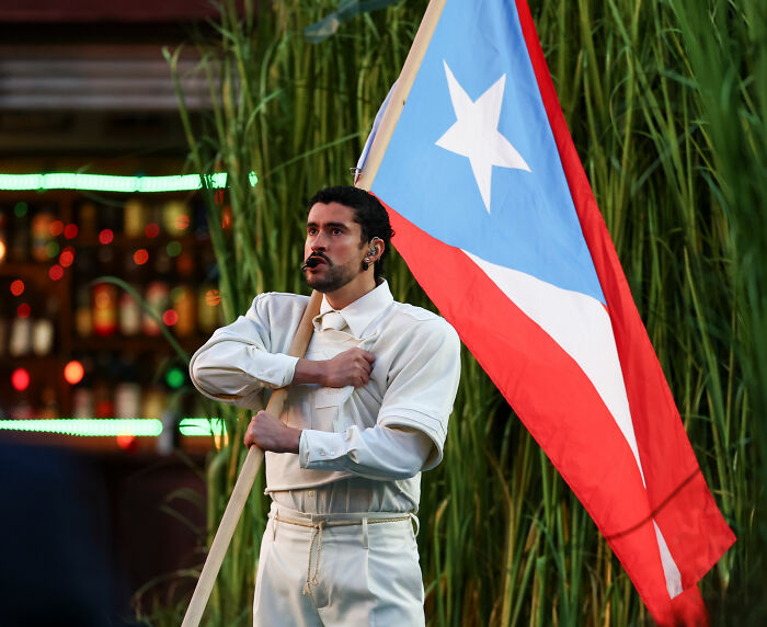 Bad Bunny holding Puerto Rican flag with a serious expression during a performance at the Super Bowl event.