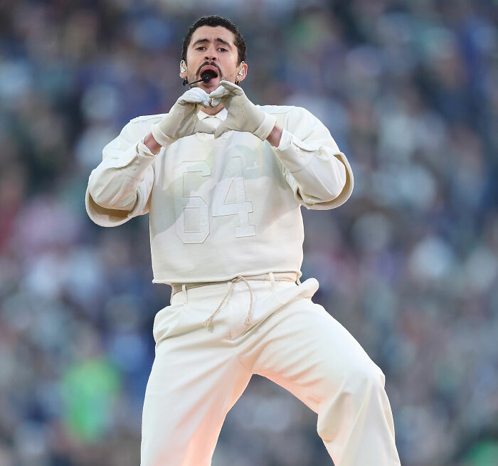 Bad Bunny performing at Super Bowl, wearing white outfit with number 64, with expressive gesture and intense focus on stage.