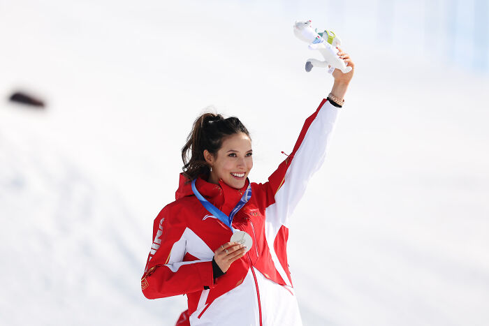 Eileen Gu in red and white jacket holding a silver medal and mascot, representing athlete in a snowy setting.
