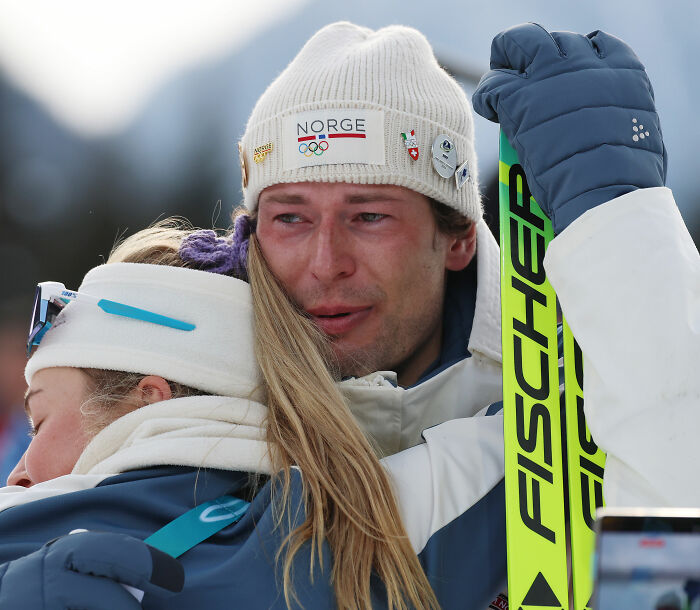 Olympian emotional while hugging girlfriend outdoors, both wearing winter sports gear, showing hurt and support.