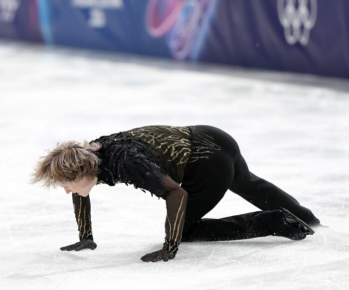 Figure skater Ilia Malinin in black and gold costume falling on ice during a competitive performance at the Olympics.