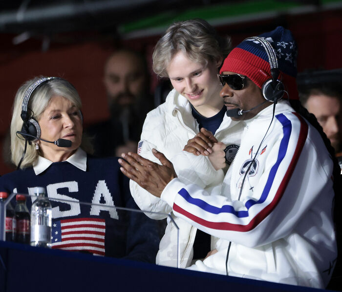NBC Olympics president wearing headset claps back while Ilia Malinin interacts with colleagues during event broadcast.