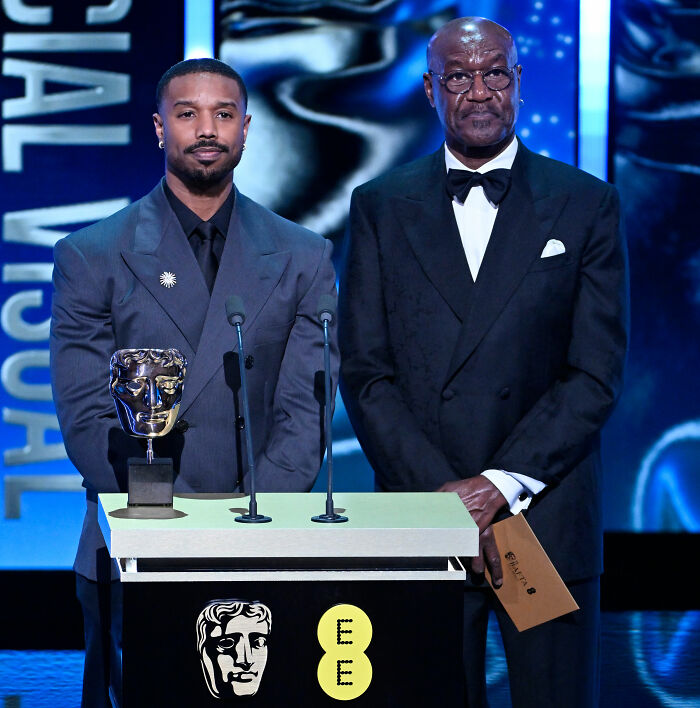 Michael B. Jordan at BAFTA ceremony with trophy, reacting after audience member incident, standing beside a man in a tuxedo.