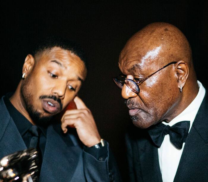 Michael B. Jordan reacting at BAFTA event with a serious expression, wearing a black suit and bow tie in dim lighting.