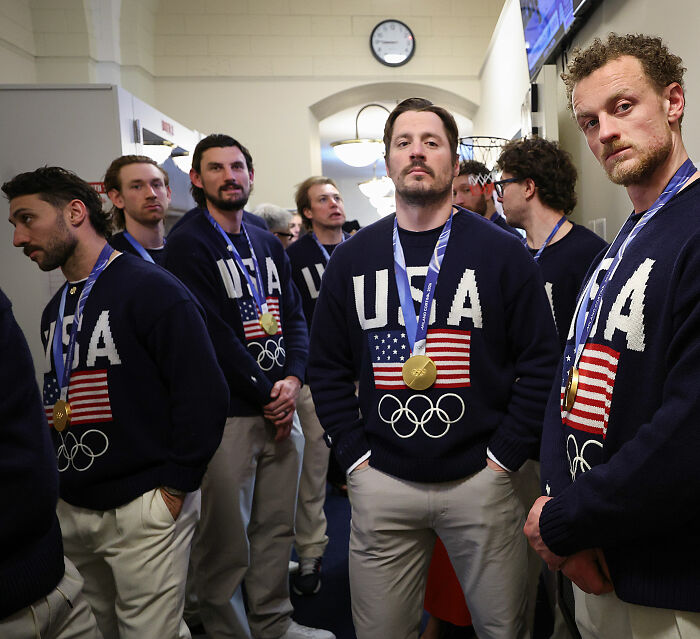 Team USA men's hockey players wearing USA sweaters and gold medals, standing indoors with serious expressions. Team USA men's hockey players wearing USA sweaters and gold medals, standing indoors with serious expressions.