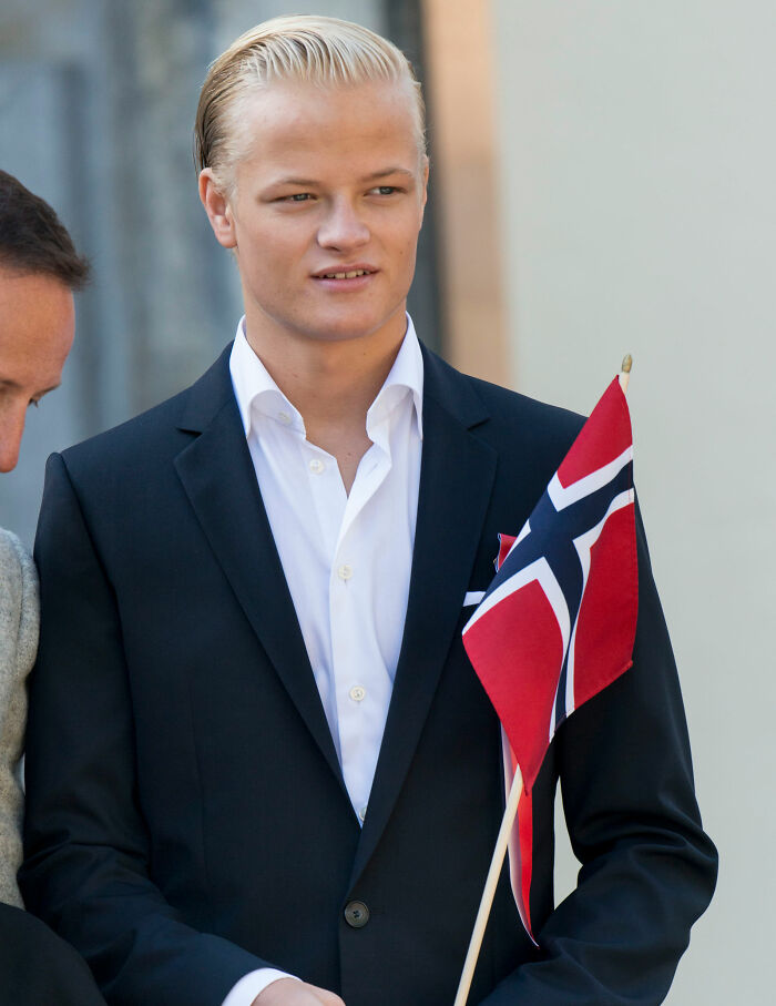 Young man holding Norway flag in a suit, representing son of Norway's crown princess in court setting.