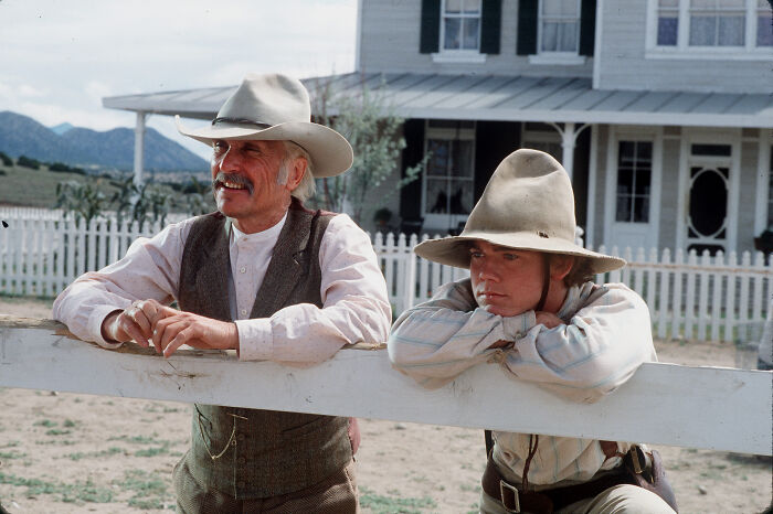 Robert Duvall in a classic western TV role wearing a cowboy hat leaning on a white fence outside a farmhouse. Robert Duvall in a classic western TV role wearing a cowboy hat leaning on a white fence outside a farmhouse.