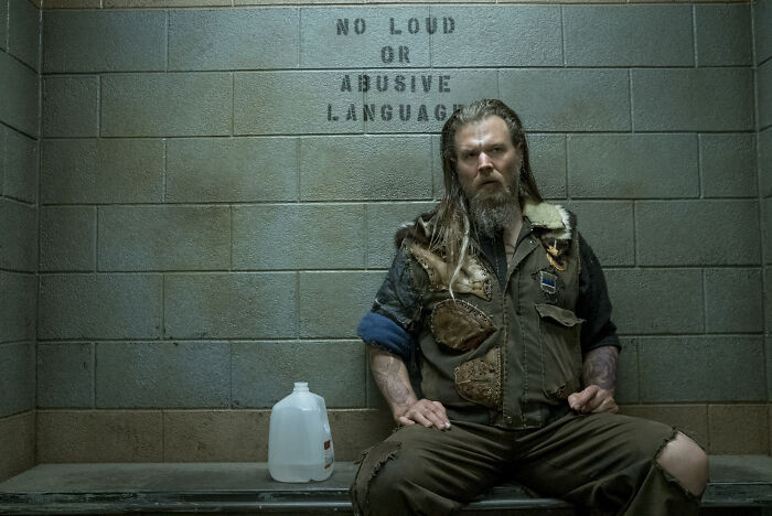 Man with long hair and beard sitting in a cell under a sign, related to God of War TV show casting Atreus.