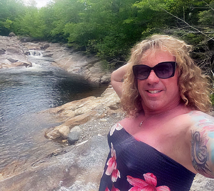 Transgender mom Roberta Esposito wearing sunglasses and floral dress by a rocky river with trees in background.