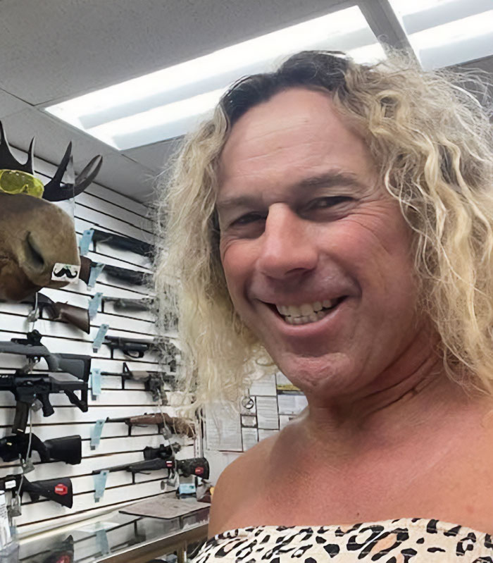 Transgender mom Roberta Esposito smiling indoors with guns displayed on the wall behind her in a store setting.