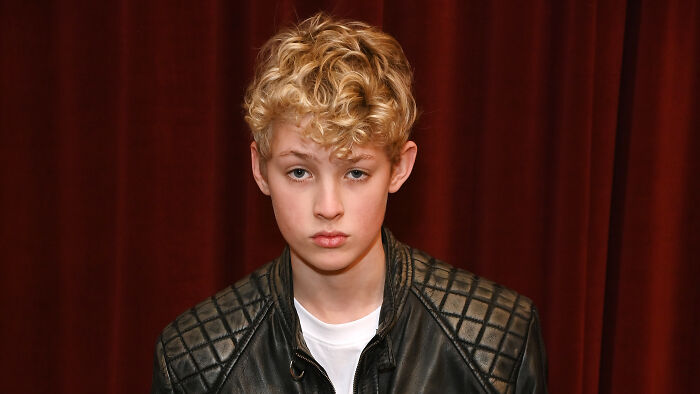 Young actor portraying Draco Malfoy in upcoming Harry Potter series wearing a black leather jacket against a dark red curtain.