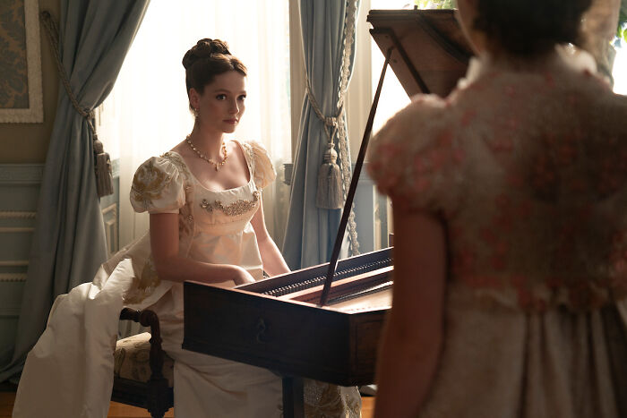 Francesca from Bridgerton dressed in period attire, sitting at a harpsichord in a softly lit room. Francesca from Bridgerton dressed in period attire, sitting at a harpsichord in a softly lit room.
