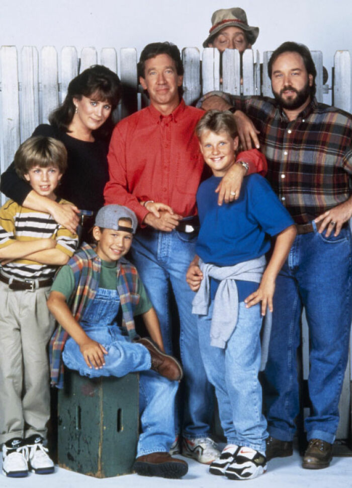 Cast of Home Improvement posing together in casual 90s clothing with a white wooden fence backdrop.