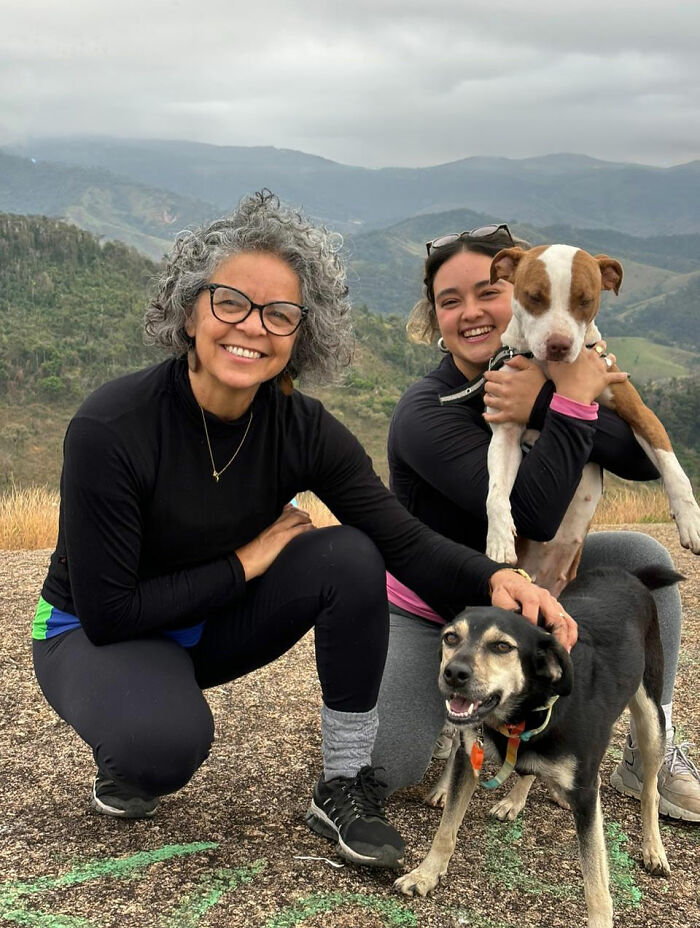 Two women outdoors smiling with a rescued pit bull and another dog in a mountainous area in Brazil.
