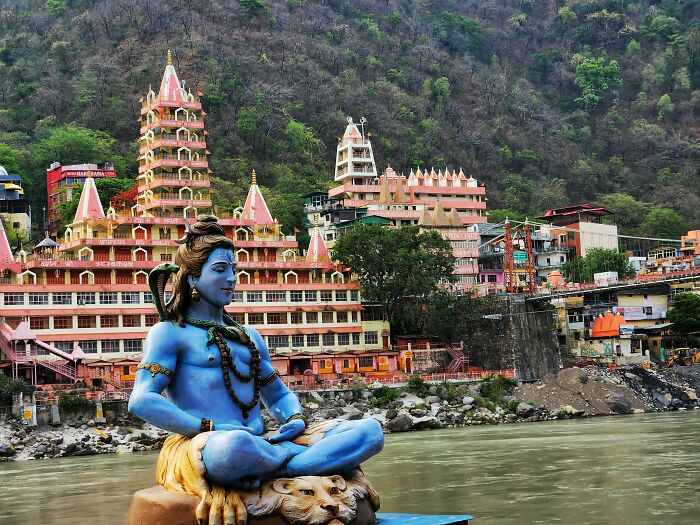 Statue of meditating Shiva by the river flows with temples in the background, highlighting spiritual tourism in India.