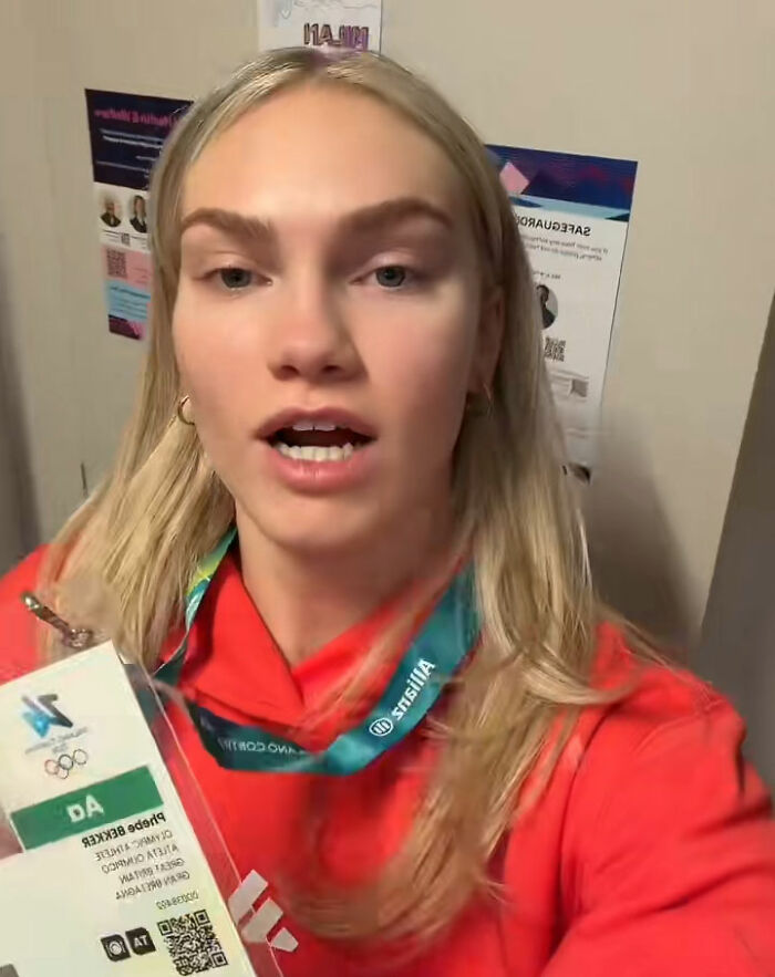 Female athlete wearing red hoodie and Winter Olympics Milan accreditation card, revealing athlete rooms after Paris beds went viral.