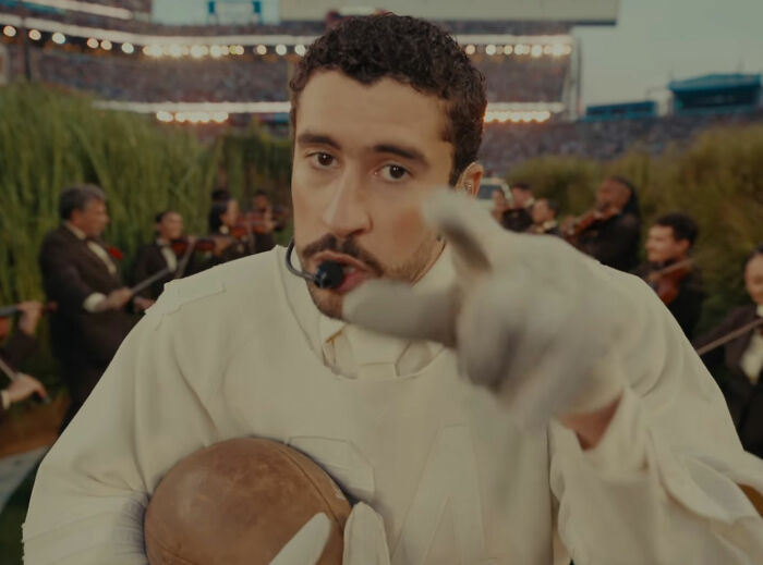 Bad Bunny performing at an outdoor event, wearing vintage sports gear and engaging with the camera closely.