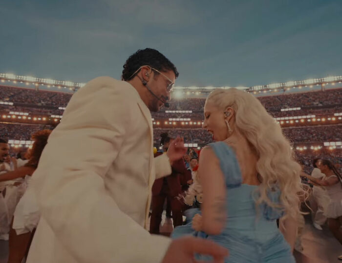 Bad Bunny exiting Super Bowl stadium looking concerned during a live performance with a female dancer in blue dress.