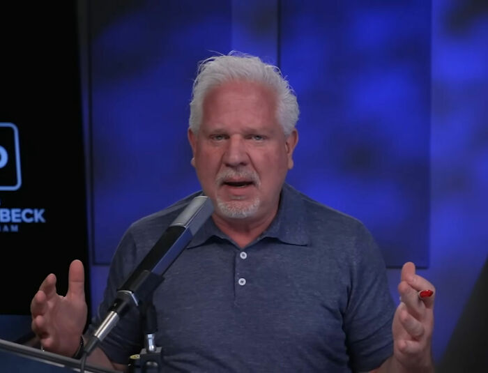 Man with white hair speaking into a microphone during a broadcast about Erika Kirk changing husband's office and TSUPA backlash.