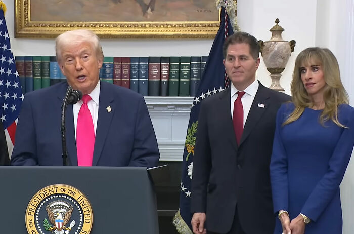Former President Trump speaking at a White House press conference with a woman exhibiting strange behavior beside him.