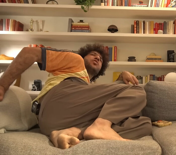Benny Blanco sitting casually on a couch in a cozy room with shelves of books and decor behind him.