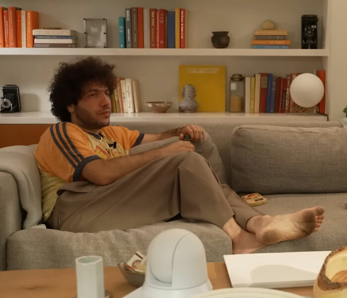 Benny Blanco sitting barefoot on a sofa in a casual home setting discussing viral interview and Selena Gomez divorce rumors.