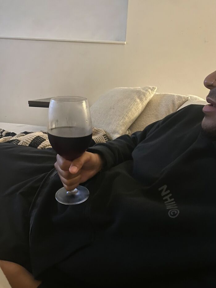 Man displaying awkward body language holding a glass of red wine while reclining on a couch with cushions behind him