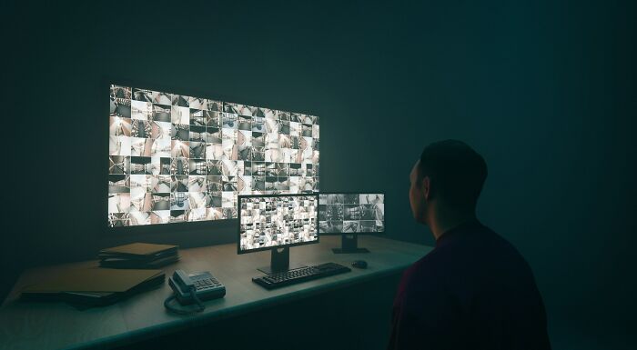 A graveyard shift worker monitoring multiple surveillance screens in a dark control room, appearing focused and alert.