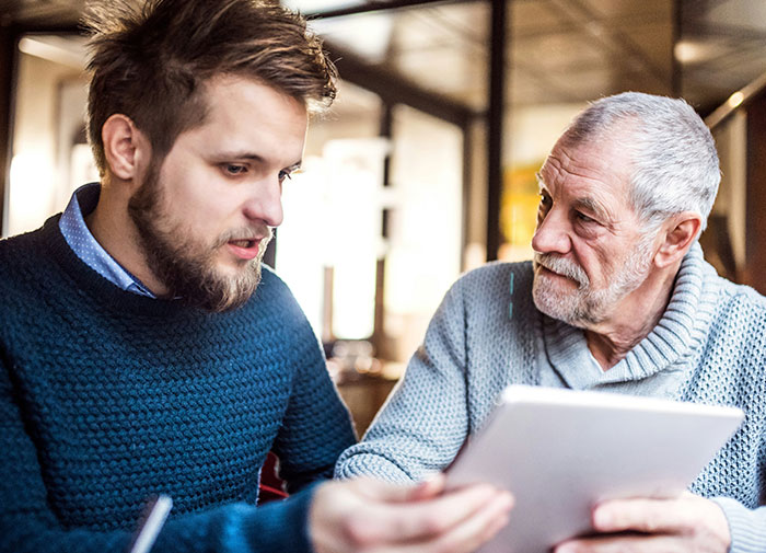 Young man and older man discussing family drama over last name while looking at a tablet in a cozy setting.