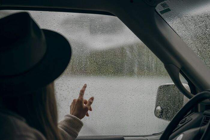 Person wearing a hat sitting in a car, pointing at raindrops on the window, capturing a thoughtful pause moment.