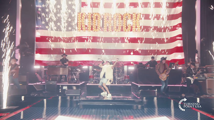 Kid Rock performing on stage with band and large American flag backdrop during alternative Super Bowl show by Turning Point USA.