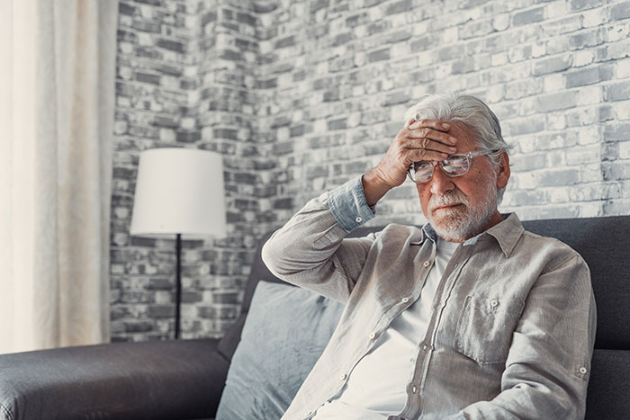 Elderly man sitting on a couch holding his forehead, illustrating stress and mental traps faced by archaeologists at job sites.