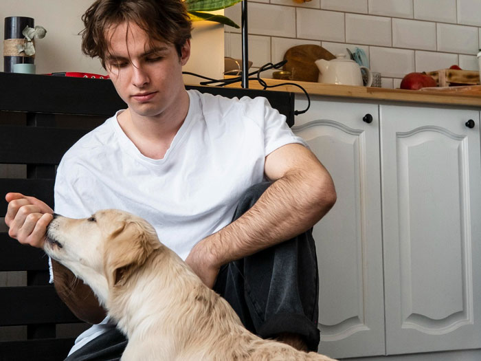 Young man sitting on the floor feeding a golden retriever dog inside a kitchen, showing pet ownership and care.