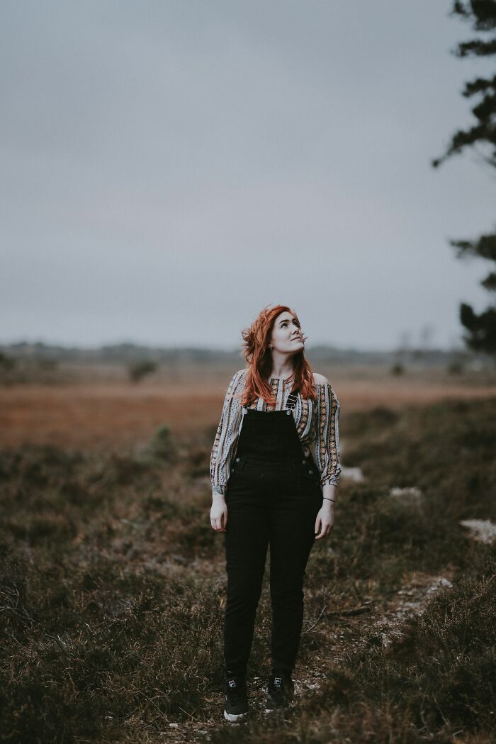 Young woman standing alone outdoors in a field, looking up with a concerned expression about immediate disaster sounds.