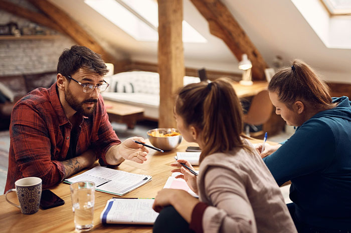 Guy refusing to pay for hotel while discussing plans with roommates in a cozy, rustic attic room setting.