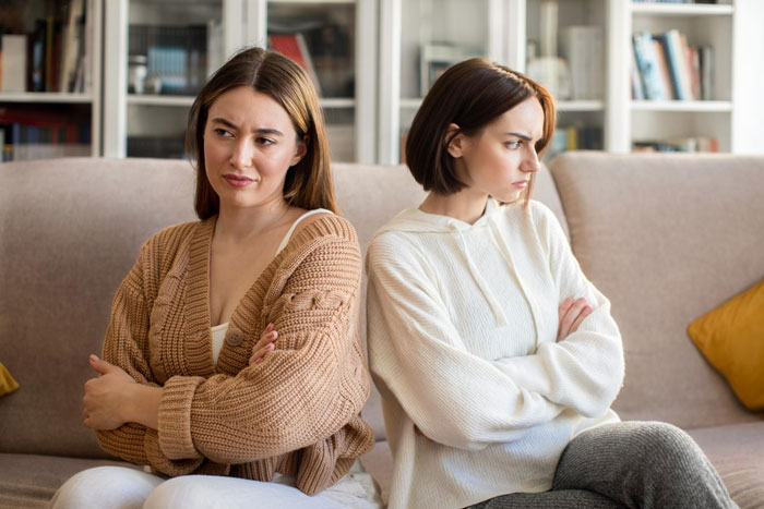 Two women sitting back to back on a couch, upset and avoiding each other after a family disagreement over spoiled child behavior.