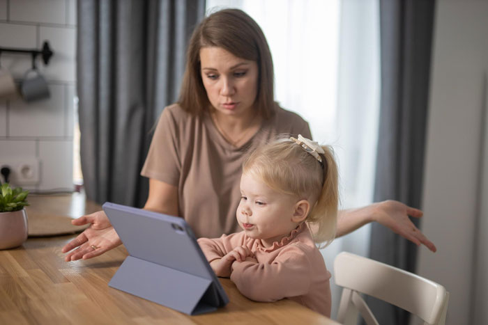 Woman looking frustrated while young girl plays on tablet, illustrating family tension over spoiled child behavior.