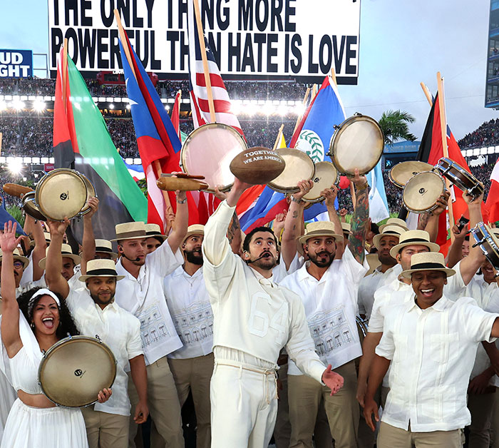 Group of Super Bowl performers in traditional attire holding drums and flags during heated debate after fans translate Bad Bunny's explicit lyrics