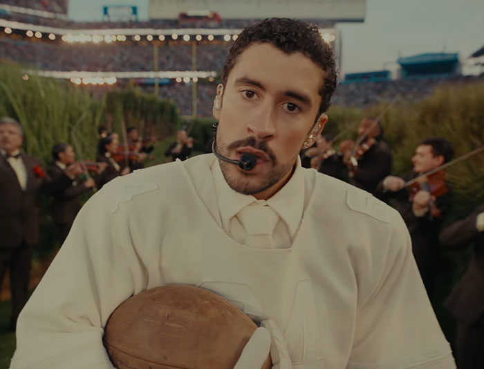 Bad Bunny wearing headset and holding football during Super Bowl performance with musicians in the background.