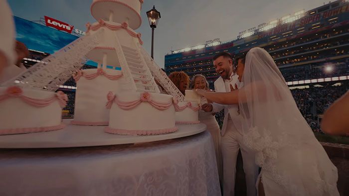 Couple in wedding attire cutting a large decorated cake during Bad Bunny's Super Bowl halftime show celebration.